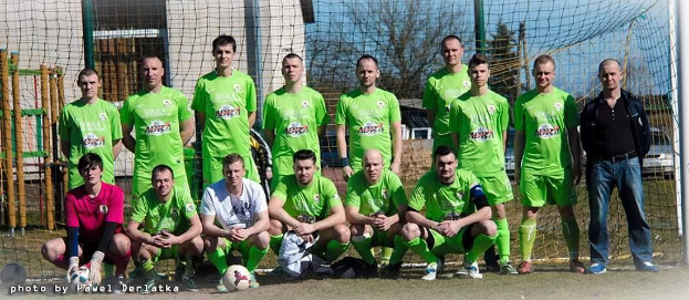 Gruppe von Männern in grünen T-Shirts auf einem Fußballfeld in der Nähe eines Torpfostens, einige in Hockstellung, mit einem Ball davor und städtischer Umgebung.