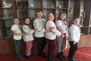 Eine Gruppe von Kindern in traditioneller ukrainischer Kleidung steht vor einem Bücherregal mit Spielzeugen, einem Skelett und anderen Gegenständen, was auf eine ukrainische Schulumgebung hinweist.
