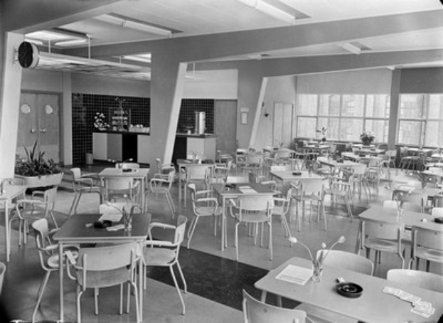 Schwarz-weißes Foto einer Cafeteria mit Tischen und Stühlen, Tellern, Besteck, Gläsern und Blumenvasen auf den Tischen sowie einer Wand mit Türen, Fenstern und Deckenleuchten im Hintergrund.