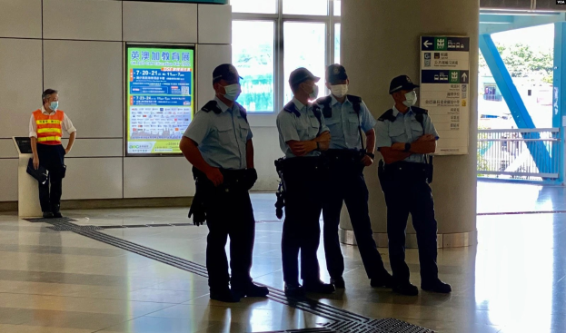 Gruppe von maskierten Polizisten vor einem Gebäude mit Glastüren, Schildern mit Haftbefehlen, einer Säule und einem Geländer, mit Bäumen und Fahrzeugen im Hintergrund.