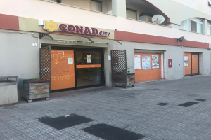 Ein Gebäude mit Fenstern, Türen und einem Schild mit der Aufschrift "Conad City" ist von Topfpflanzen, einem Metallzaun und einem bewölkten Himmel umgeben, mit Papieren an der Wand.