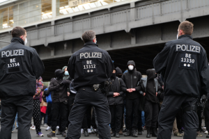 Polizeibeamte in schwarzen Uniformen und Masken stehen vor einer Menge auf einer Demonstration, mit einer Brücke und einem Gebäude im Hintergrund.