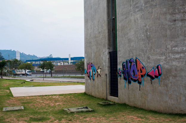 Betonschätze mit Graffiti, umgeben von Gras, Bäumen, Masten, Fahrzeugen auf der Straße, anderen Gebäuden, Hügeln und einem klaren blauen Himmel.