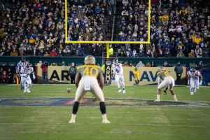 Fußballspieler in Sportkleidung und Helmen konkurrieren auf einem Feld mit Zuschauern im Hintergrund, umgeben von Stadionstrukturen und Werbetafeln.