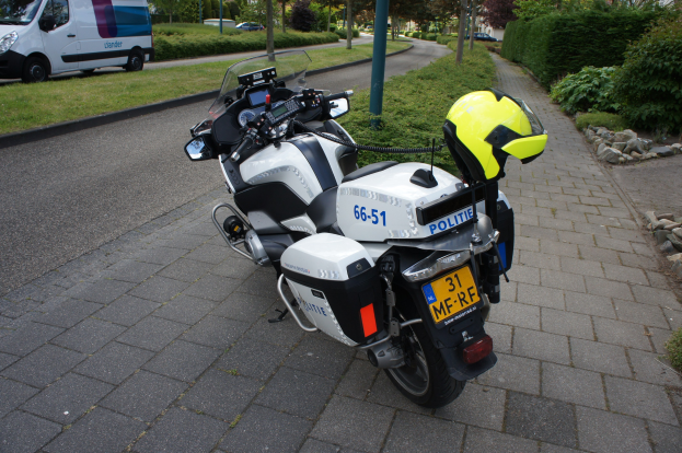 Ein polizeiliches Motorrad am Straßenrand mit einem Helm darauf, umgeben von Bäumen, Pfählen, Gebäuden, Pflanzen, Gras, Steinen und anderen Fahrzeugen.