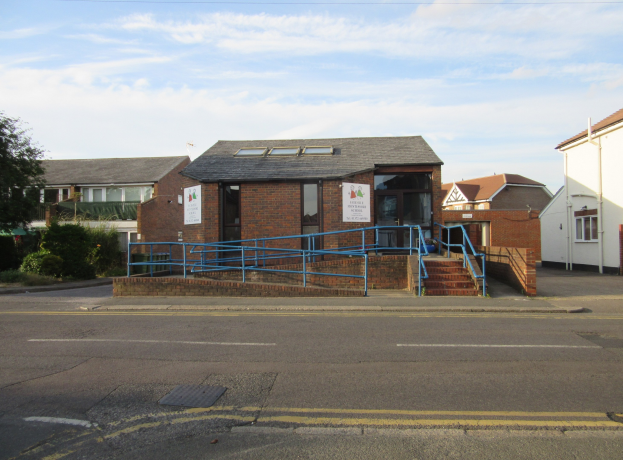 Ein Schulgebäude mit einer blauen Begrenzung davor, umgeben von Häusern, Treppen mit Begrenzungen, Informationsschildern, Pflanzen, Bäumen und einem bewölkten Himmel.