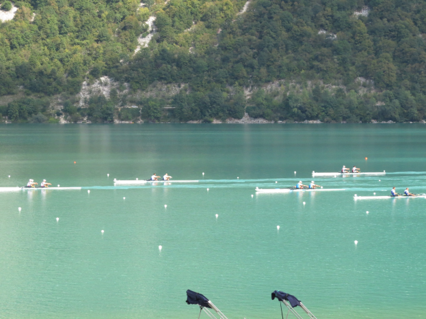 Eine Gruppe von Menschen bei einem Ruderwettbewerb auf einem Gewässer mit Bäumen und einem Hügel im Hintergrund, mit Kleidung auf Ständen am Boden.