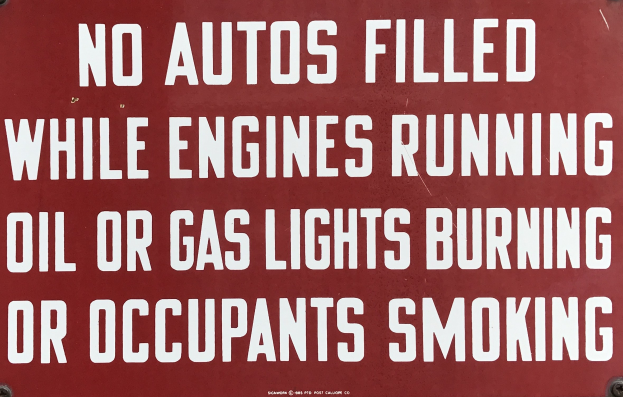 Ein rotes Schild mit weißer Schrift, das davor warnt, Autos während des Laufens des Motors oder mit Öl, Gas oder rauchenden Insassen aufzufüllen.