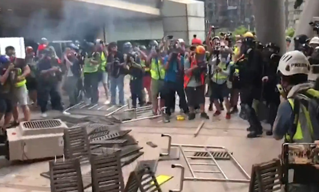 Eine Gruppe von Menschen in Helmen und Jacken steht in der Nähe eines rauchenden Gebäudes, einige halten Kameras, mit verstreuten Metallobjekten und Stadtpfeilern im Hintergrund.