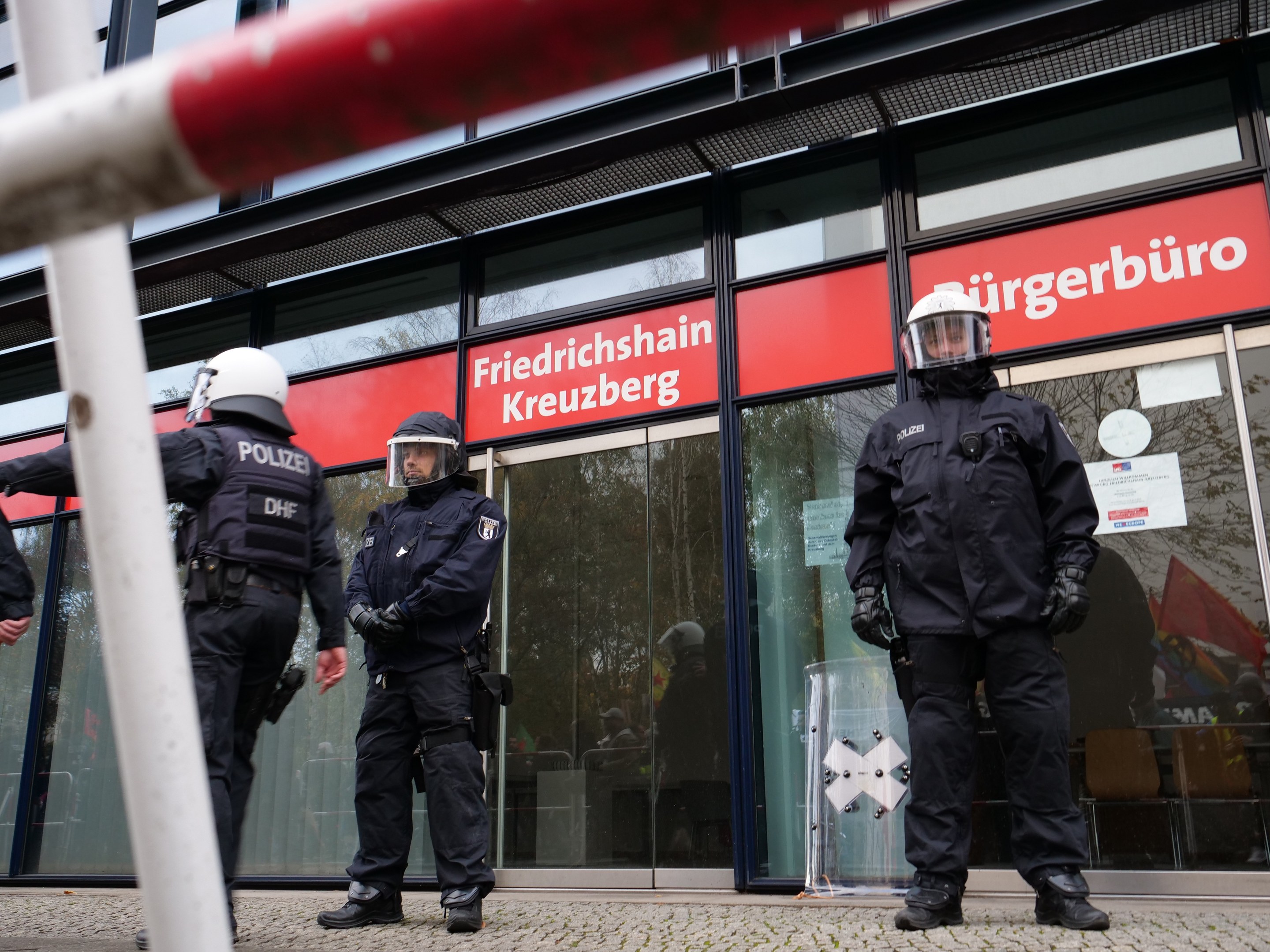 Polizisten in Helmen und Handschuhen vor einem Gebäude mit Glaswänden und -türen, neben einem Pfahl, mit "Friedrichshain Kreuzberg"-Text auf Schildern im Hintergrund.