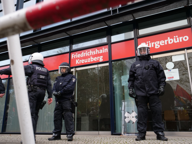 Polizisten in Helmen und Handschuhen vor einem Gebäude mit Glaswänden und -türen, neben einem Pfahl, mit "Friedrichshain Kreuzberg"-Text auf Schildern im Hintergrund.