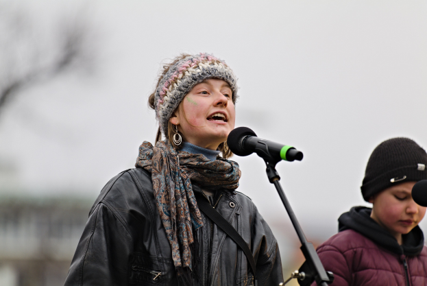 Eine Frau in einer schwarzen Jacke, einem Schal und einer Mütze singt in ein Mikrofon auf der Bühne, neben einer Person in einer maronenfarbenen Jacke und einer schwarzen Mütze, während der unscharfe Hintergrund einen träumerischen Effekt erzeugt.