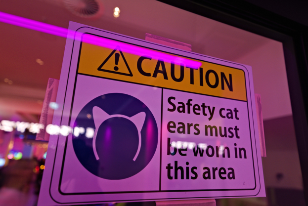 Hellgelbes Warnschild in einem Ladengeschäft mit der Aufschrift "Achtung: Sicherheits-Katzenohren müssen in diesem Bereich getragen werden." vor einem unscharfen Hintergrund.