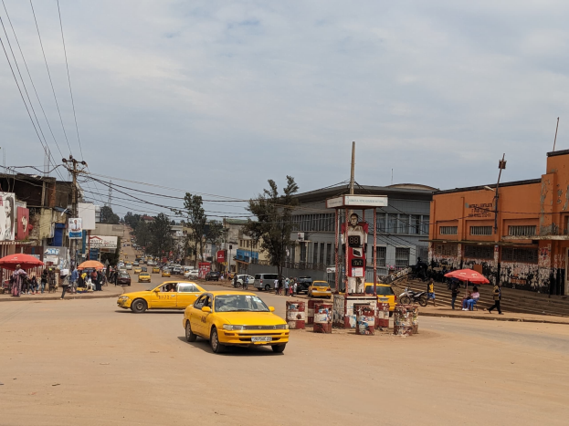 Ein gelbes Taxi fährt eine belebte Stadtstraße entlang, gesäumt von Gebäuden, während Fußgänger mit Regenschirmen auf dem Gehweg darunter gehen und Wolken den Himmel bedecken.