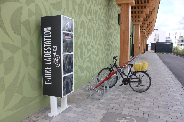Ein Fahrrad, das vor einem Gebäude mit einem "E-Bike-Ladestation"-Schild geparkt ist, mit einem Rad im Vordergrund und Gebäuden, Bäumen, Pflanzen und einem klaren blauen Himmel im Hintergrund.
