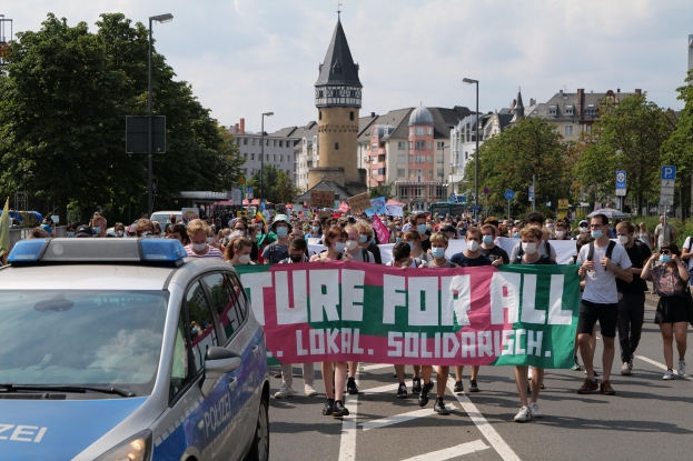 Eine große Gruppe von Menschen marschiert mit einem Banner mit der Aufschrift "Lokale Solidarität" eine Straße entlang, wobei ein Polizeiwagen am Straßenrand geparkt ist und Gebäude, Bäume und Straßenelemente im Hintergrund zu sehen sind.