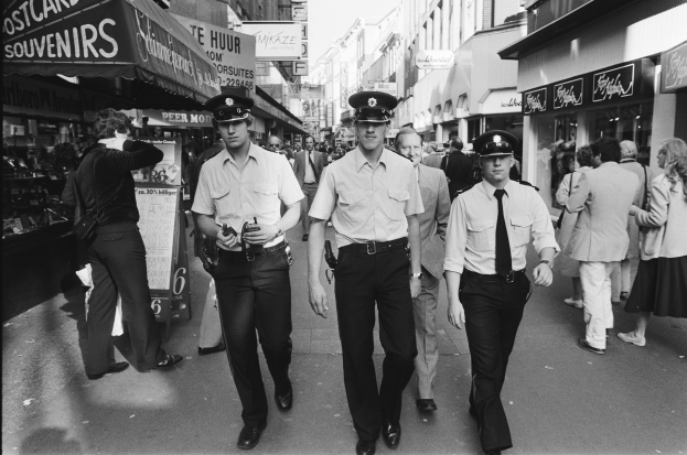 Drei Polizeibeamte in Uniform gehen eine belebte Straße mit hohen Gebäuden und Geschäften entlang, mit Passanten und einem sichtbaren Himmel im Hintergrund.