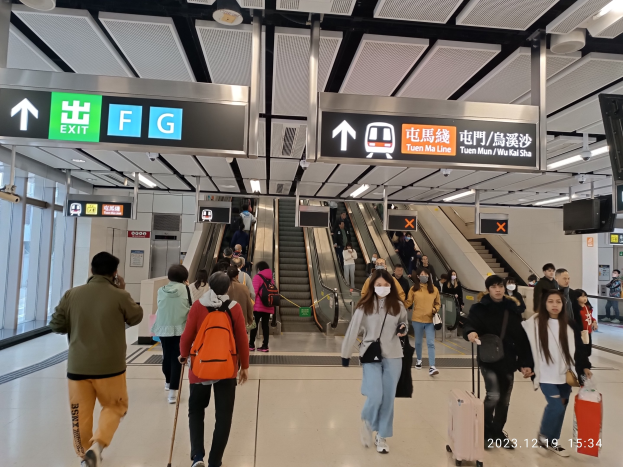 Menschen mit Gepäck durch einen geschlossenen Flughafen mit Rolltreppen, Informationsanzeigen, Deckenleuchten und Glaswänden im Hintergrund.
