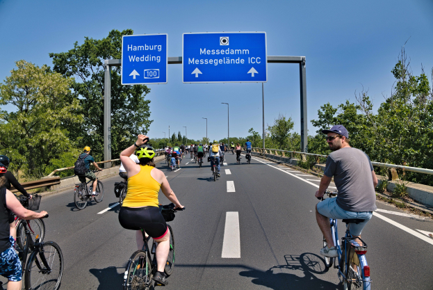 Gruppe von Radfahrern mit Helmen auf einer Straße mit einer Begrenzung auf der linken Seite und Bäumen auf der rechten Seite, Laternen im Hintergrund, unter einem klaren blauen Himmel, mit einem Schild oben, das eine Radtour in Hamburg anzeigt.