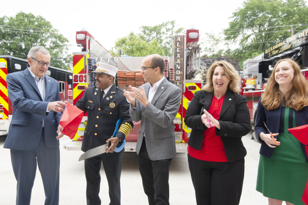 Group of people clapping and smiling at a ribbon cutting ceremony for the Alexandria Fire Department, with two holding scissors and a red ribbon in front of a fire truck.