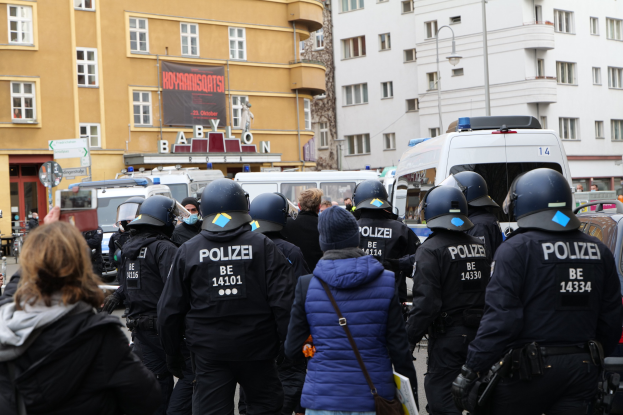 Polizisten in Uniform vor einer Menge von Demonstranten während einer Demonstration in Berlin, mit Fahrzeugen, Gebäuden und einem Kameramann im Hintergrund.