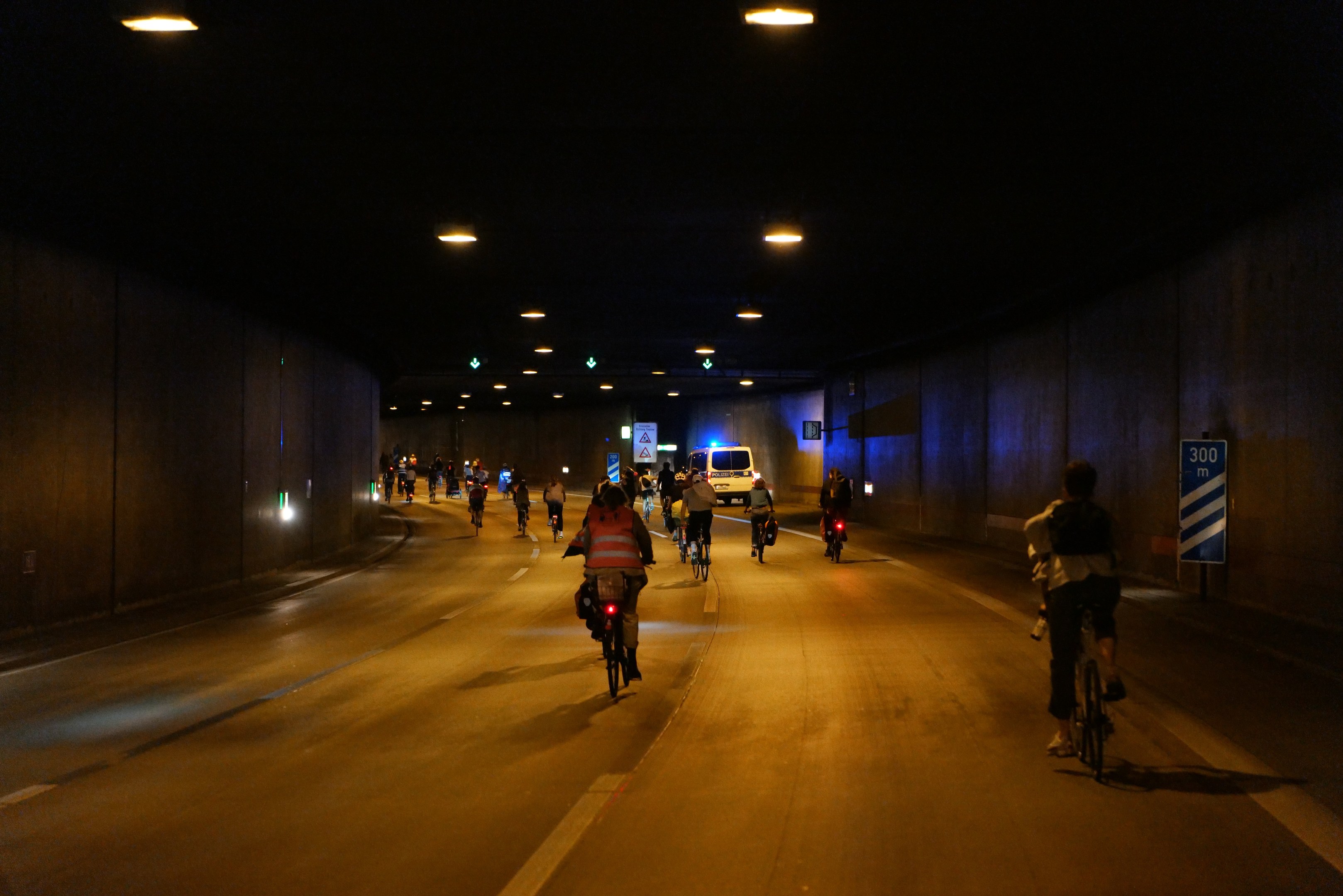 Eine Gruppe von Menschen, die nachts auf Fahrrädern eine Straße entlangfahren, mit über ihnen leuchtenden Lichtern, flankiert von Wänden und einem im Hintergrund sichtbaren Fahrzeug.