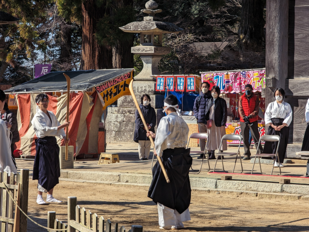 Eine Gruppe von Menschen in formeller Kleidung, einige tragen Masken und halten Stöcke, versammelt sich vor einem traditionellen japanischen Gebäude während einer Zeremonie in Kyoto, mit Stühlen, einem Zelt, Bannern, Bäumen, einem hölzernen Zaun und einem klaren blauen Himmel.