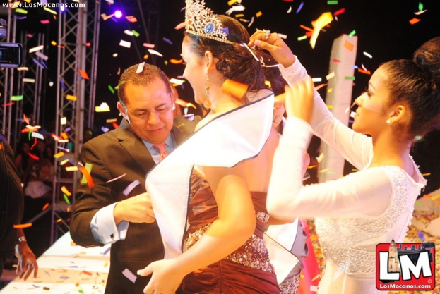 Ein Mädchen auf der Bühne bei einer Miss-World-Show mit einer Krone, mit einem anderen Mädchen, das sie von hinten anpasst, und einem Mann in einem schwarzen Anzug, der vor ihr die Hand schüttelt, vor einem dunklen Hintergrund mit Metallrahmen.