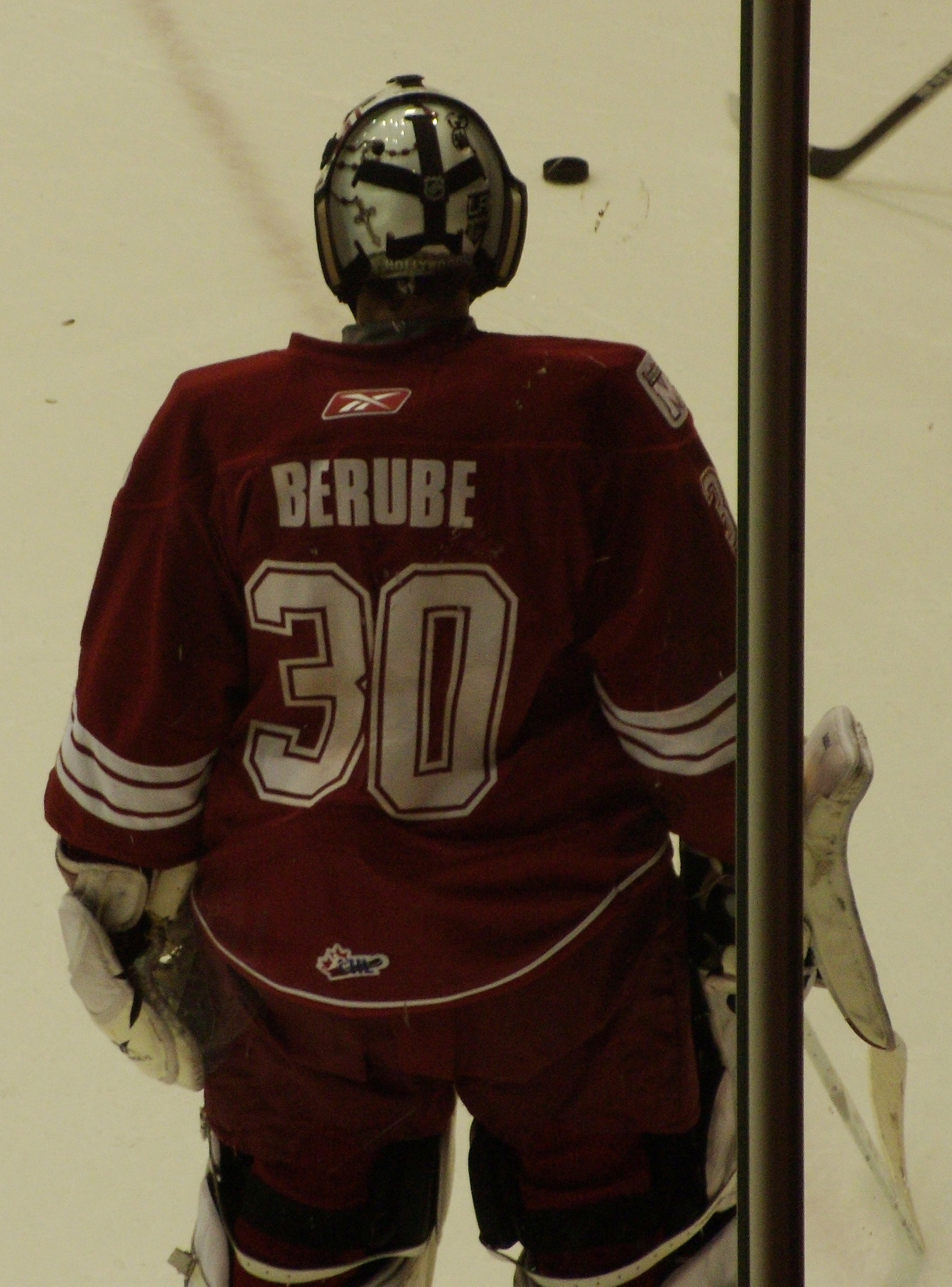 Ein Hockey-Spieler in Helm, Handschuhen und nummeriertem Trikot steht auf dem Eis vor einem Tor mit einem Pfosten rechts daneben.