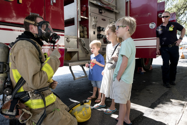 Feuerwehrmann mit Gasmask in Gespräch mit Kindern vor einem Feuerwehrauto mit Bäumen und blauem Himmel im Hintergrund.