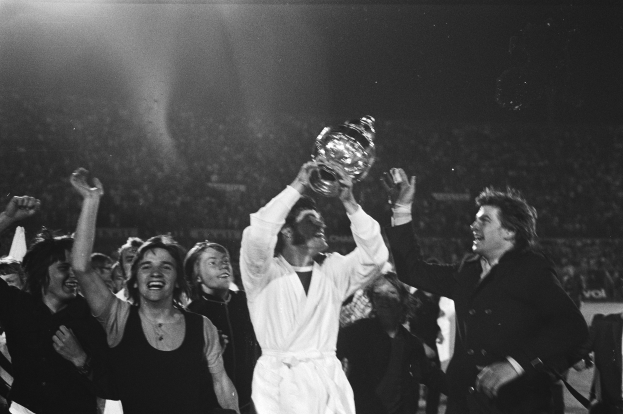 Eine Gruppe von Menschen feiert mit einem Pokal vor einer jubelnden Menge, wobei die zentrale Figur den Pokal in Schwarzweiß hochhält.
