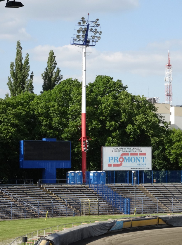 Rennstrecke beleuchtet von oben, umgeben von Gras, Geländern, Stufen, einem Lichtmast, einem Schild, Bäumen, Gebäuden, Türmen und einem bewölkten Himmel.