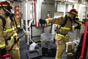 Drei Feuerwehrleute in Schutzausrüstung stehen zusammen in einem Raum mit einem Feuerlöscher links, einer Maschine rechts und verstreuten Gegenständen auf dem Boden.
