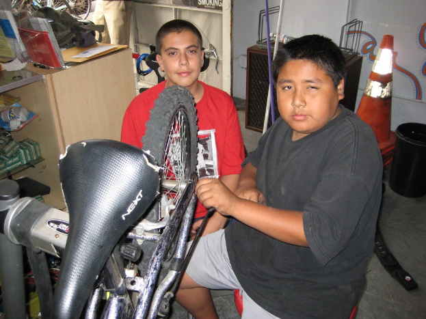 Zwei Personen reparieren ein Fahrrad in der Mitte; ein Schreibtisch mit Ordnern, Taschen und Kisten steht links, während Fahrräder, Ersatzteile und andere Gegenstände den Hintergrund füllen.