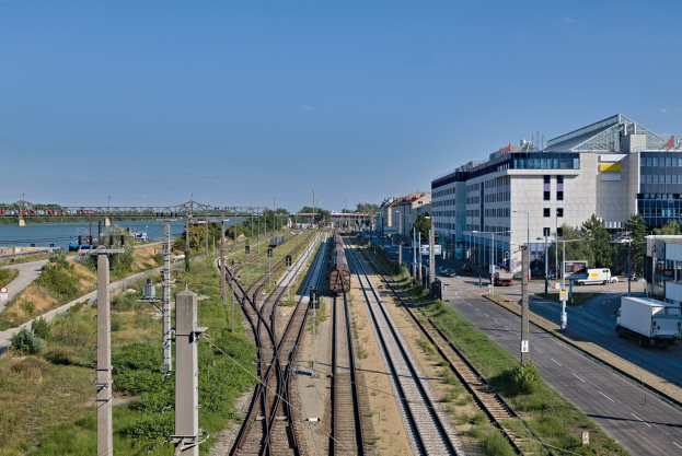 Ein Zug fährt auf Schienen neben einer Stadtlandschaft mit Gebäuden, Fahrzeugen, Bäumen und einer Brücke unter einem bewölkten Himmel.