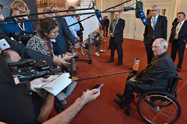 Ein Mann im Rollstuhl umgeben von Reportern mit Kameras, Mikrofonen und Handys sowie einem Banner und Scheinwerfern im Hintergrund.