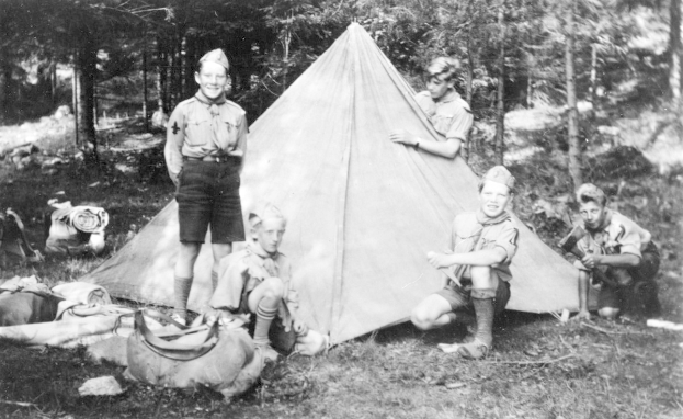 Schwarzes und weißes Foto von Pfadfindern in Uniform, die sich um ein Zelt in einem bewaldeten Gebiet versammelt haben, einige sitzen auf dem Boden, während andere stehen, mit verstreuten Taschen und Equipment.
