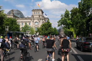 Eine Gruppe von Menschen, die auf Fahrr√§dern eine von B√§umen gesäumte Straße in Berlin, Deutschland, entlangfahren, mit Gebäuden auf beiden Seiten, einer Bushaltestelle auf der rechten Seite und einer Flagge, die auf einem der Gebäude weht, unter einem bewölkten Himmel.