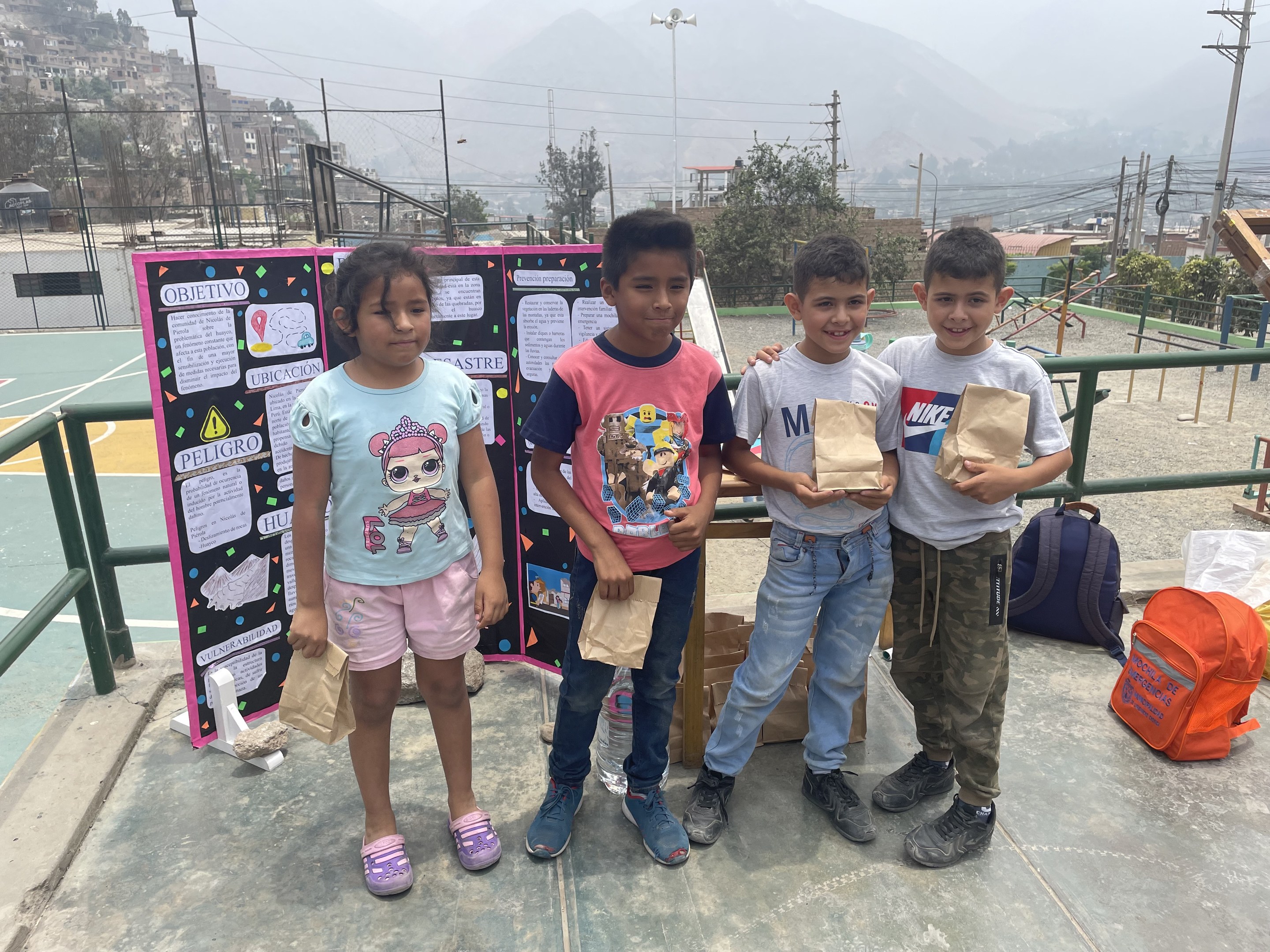 Gruppe von Kindern auf einem Basketballfeld mit Papierbeuteln in den Händen, mit Beuteln und Gegenständen rechts und einer Tafel mit Text links, vor einer Außenkulisse mit Zaun, Pfosten und Gebäuden.