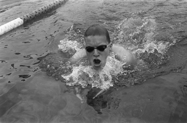 Ein Mann schwimmt in einem Pool mit offenem Mund und trägt eine Schwimmbrille, umgeben von Bahnmarkierungen, in einem Schwarz-Weiß-Bild.