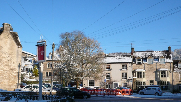 Schneebedeckte Fahrzeuge auf einer Straße mit Häusern, Bäumen, Strommasten und Drähten im Hintergrund, alles von Schnee bedeckt.