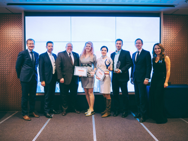 Gruppe von lächelnden Menschen mit Urkunden, die vor einem Bildschirm mit einem Tisch und einem Objekt im Hintergrund stehen, Gewinner der Internationalen Geschäftsauszeichnungen 2018.