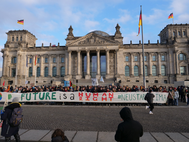 Gruppe von Menschen mit einem Banner mit der Aufschrift "Zukunft ist ein Mensch Neustar ima" vor dem Reichstaggebäude in Berlin, Deutschland, mit seinen architektonischen Details und umgeben von Flaggen, die unter einem bewölkten Himmel sichtbar sind.