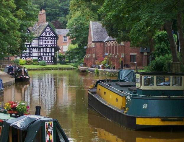 Kanal mit mehreren Booten, die an seinen grasbewachsenen, baumbestandenen Ufern liegen, mit Gebäuden im Hintergrund.
