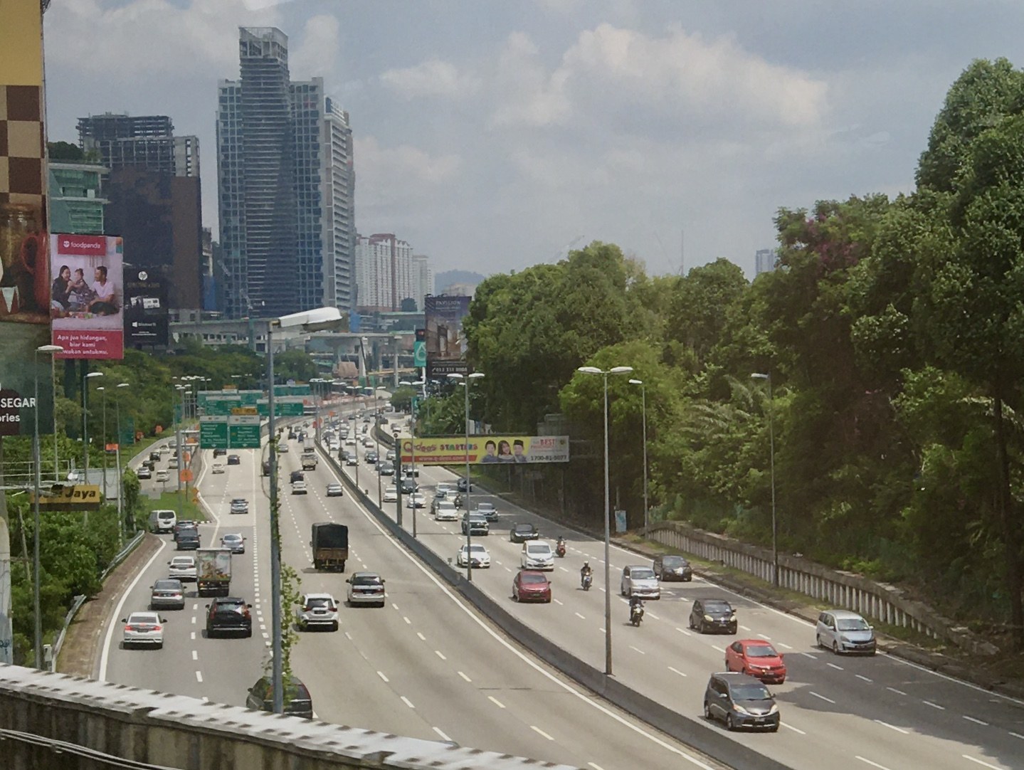 Eine vielbefahrene Autobahn mit hohen Gebäuden, Straßenlaternen, Lichtern, Werbetafeln, Bäumen und einem bewölkten Himmel.