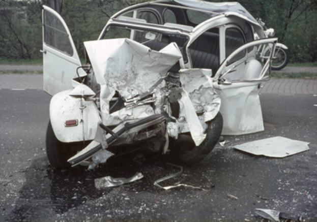 Ein verunglücktes Auto mit schwerer Frontschäden am Straßenrand, mit Bäumen, einem Gebäude und einem weiteren Fahrzeug im Hintergrund.