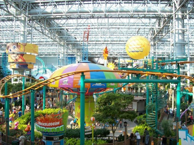 Indoor-Wasserpark mit Achterbahn, umgeben von Menschen, Bäumen, Pflanzen und Deckenleuchten mit Stangen.