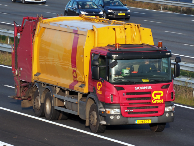 Ein gelb-roter Müllwagen fährt auf einer Autobahn, umgeben von anderen Fahrzeugen, mit einer Begränzung und Gras auf beiden Seiten der Straße.