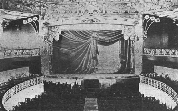 Schwarzes und weißes Foto des Auditoriums des Theatre Royal in London, das eine Bühne mit einem Vorhang, Geländern und ornamentalen Wanddesigns zeigt, mit einigen sitzenden Zuschauern und Text unten.