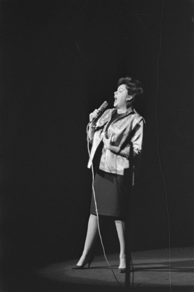 Eine Frau in einem schwarzen und weißen Outfit aus den 1950er-Jahren singt leidenschaftlich auf einer von einem Scheinwerfer beleuchteten Bühne und hält ein Mikrofon.